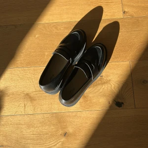 Svarta loafers i skinn från Alley - Stilrena svarta nya loafers från Alley i glansigt skinn. Klassisk penny loafer-design med rund tå och platt sula. Perfekta för dig som vill ha en tidlös och clean look till vardagsoutfiten.