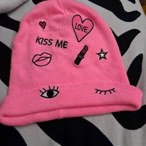 Rosa mössa med broderade motiv - Cool rosa mössa med broderade detaljer som hjärta, stjärna, ögon, läppar och texten 'KISS ME' och 'LOVE'. Mössan är stickad och har en uppvikt kant med ögonmotiv. Den har en fläck som syns när man viker ner den, men borde gå att tvätta bort. Fungerar för barn och vuxna.