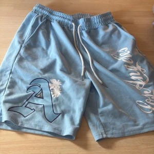 Blå shorts med tryck och snörning - Snygga ljusblå shorts med vit snörning i midjan och tryck med bokstaven A och palmträd på ena benet. Mjukt material och avslappnad passform, perfekta för sommardagar. Stora fickor på sidorna och elastisk midja för extra komfort.