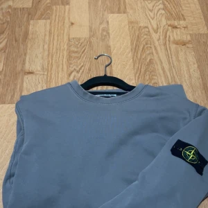Grå sweatshirt från Stone Island - Säljer en stilren grå sweatshirt från Stone Island med klassisk rund hals och ikonisk logotyp-patch på vänster ärm. Tröjan har en enkel design och är tillverkad i mjuk bomull, perfekt för en clean och avslappnad look.