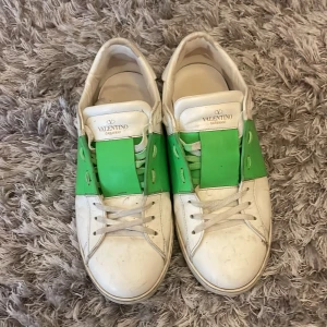 Valentino Garavani sneakers med grönt band - Snygga vita Valentino Garavani sneakers med ett grönt band över vristen och coola nitar på hälen. Skorna har snörning och är tillverkade i skinn. Skorna är köpta på The Grand archive och är 100% äkta