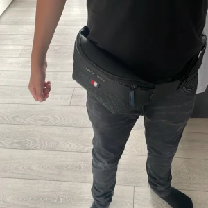 Svart magväska Tommy Hilfiger - Säljer en svart magväska från Tommy Hilfiger med präglat mönster och klassisk logga framtill. Väskan har justerbart band med broderad text och dragkedja upptill. Perfekt för att bära det viktigaste och ge outfiten en clean vibe.