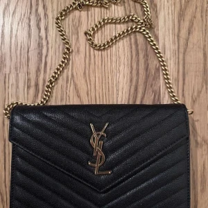 Svart YSL kuvertväska i skinn - Säljer en svart kuvertväska från Saint Laurent med klassiskt YSL-emblem i guld framtill. Väskan har quiltad design, kedjerem i guld och är tillverkad i skinn. Invändigt finns flera kortfack och ett fack med dragkedja. Perfekt för att bära det viktigaste med stil.