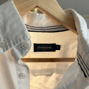 Vit skjorta slim fit från Dressmann - Klassisk vit skjorta från Dressmann i slim fit-modell. Skjortan har lång ärm, bröstficka och knappar framtill. Perfekt för en clean och stilren look. Tillverkad i mjuk bomull som känns skön mot huden.