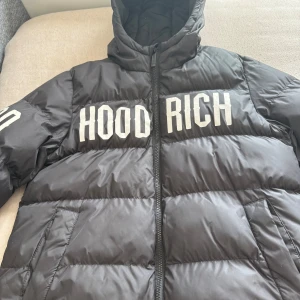 Svart pufferjacka från Hoodrich - Svart pufferjacka från Hoodrich med stor vit logga på både bröstet och ryggen. Jackan har huva, dragkedja framtill och två sidofickor. Perfekt för kalla dagar när du vill ha en streetstyle-look.