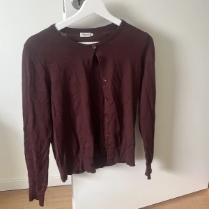 Mörkbrun kofta från Filippa K XS - En klassisk mörkbrun kofta från Filippa K i storlek XS. Koftan har rund hals, långa ärmar och knäppning framtill med matchande knappar. Tillverkad i mjukt, tunt stickat material som känns skönt mot huden. Perfekt för lager-på-lager och enkel att styla.