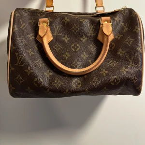 Louis Vuitton monogram handväska - Säljer en klassisk Louis Vuitton Speedy handväska i brunt skinn med ikoniskt monogrammönster. Väskan har två handtag i ljust läder, guldfärgade metalldetaljer och dragkedja upptill. Perfekt storlek för vardagens essentials och en riktig statement piece.