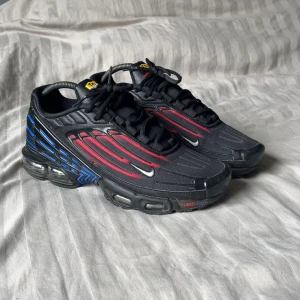 Nike Tuned Air sneakers svart/röd/blå - Nike Tuned Air sneakers med svart mesh och syntet, röda och blå detaljer längs sidorna och sulan. Snygg futuristisk design med synlig luftdämpning och små silvriga swoosh-loggor. Skorna har snörning och en chunky platt sula som ger streetkänsla.