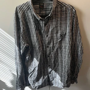 Randig skjorta i bomull Premium Cotton - Snygg långärmad skjorta från Premium Cotton i svart, vit och grått randigt mönster. Skjortan är tillverkad i mjuk bomull och har klassisk krage, knäppning framtill och bröstficka. Perfekt för dig som gillar stilrena och tidlösa plagg.