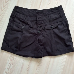 Svarta högmidjade shorts med knappar - Snygga svarta shorts med hög midja och flera dekorativa knappar framtill. Shortsen har fickor med lock och knappdetaljer både fram och bak samt uppvikta benslut. Tillverkade i ett mjukt bomullstyg som känns skönt mot huden. Perfekta för en stilren och chill look.