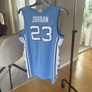 Äkta Jordan North Carolina basketlinne  - Säljer ett ikoniskt North Carolina basketlinne från Jordan med nummer 23 och namnet Jordan på ryggen. Linnet är ljusblått med vita och blå detaljer längs sidorna, meshmaterial för extra andningsförmåga och klassisk v-ringning. Perfekt för dig som älskar basket och streetwear.