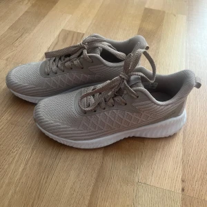 Beige sneakers från Exani - Snygga beige sneakers från Exani med stickad mesh-ovandel och vita, vågformade sulor. Skorna har snörning och diskret mönster på sidan för en sportig vibe. Perfekta för dig som gillar stilrena och bekväma skor till vardags eller träning.