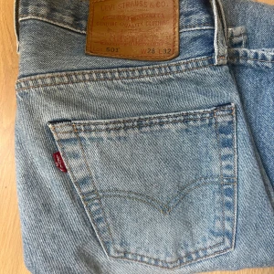 Levi's 501 ljusblå jeans W28 L32 - Klassiska Levi's 501 jeans i ljusblå denim med raka ben och normal passform. Byxorna har fem fickor, knappgylf och den ikoniska läderpatchen bak i midjan. Perfekta för en avslappnad och tidlös look.