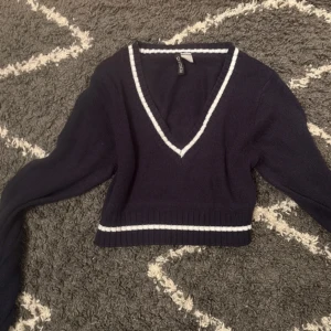Mörkblå croppad stickad tröja H&M - Snygg mörkblå croppad stickad tröja från H&M Divided med vita ränder vid v-ringningen och nederkanten. Tröjan har lång ärm och ribbade detaljer, perfekt för lager-på-lager och en sportig vibe.