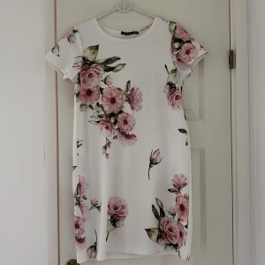 Vit blommig kortklänning - Superfin vit kortklänning med stora rosa blommor och gröna blad. Klänningen har rund halsringning och korta ärmar, perfekt för sommarens alla tillfällen. Lätt och bekväm passform som känns fräsch och romantisk.