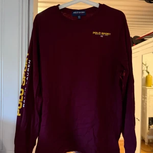 Vinröd långärmad Polo Sport tröja - Snygg vinröd långärmad tröja från Polo Sport Ralph Lauren med gul logga på bröstet och längs ena ärmen. Klassisk rund halsringning och relaxed fit. Perfekt för dig som gillar streetwear och vill sticka ut med en ikonisk look. Barn storlek i XL passar S/M