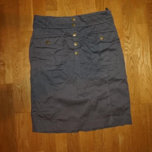 Grön kortkjol med fickor och knappar - Snygg grön kortkjol med stora fickor framtill och coola guldfärgade knappar längs mitten. Kjolen har en rak passform och är gjord i bomull, vilket gör den både bekväm och lätt att matcha med olika outfits.