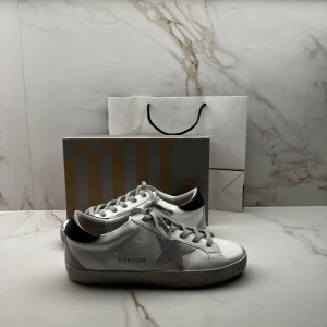 Golden Goose vita sneakers med stjärna - Säljer ett par riktigt feta vita Golden Goose sneakers med grå mockadetaljer och ikonisk stjärna på sidan. Grå snörning, svart häl och platt sula. Snygg och stilren modell som passar perfekt till grisch och casual outfits.