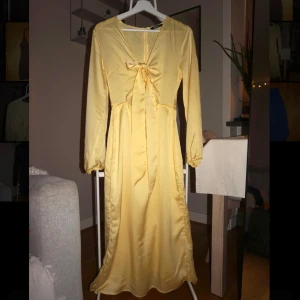 Gul långklänning från Gina Tricot - Lång gul klänning i satinliknande material från Gina Tricot. Klänningen har v-ringning, lång ärm och en söt rosett framtill. Den har en smickrande passform och dragkedja i ryggen. Perfekt för dig som vill sticka ut med en ljus och fräsch look.