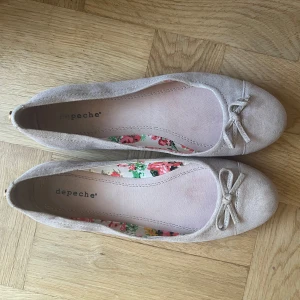 ballerinaskor  - Söta beige ballerinaskor från Depeche i mocka med rund tå och liten rosett framtill. Insidan har ett blommigt tygfoder som ger en extra touch. Skorna har platt sula och en diskret gulddetalj bak.