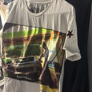 Vit Bastille t-shirt med tryck - Vit t-shirt från Bastille med stort färgglatt tryck framtill som visar en bensinmack och bil, samt en svartvit tiger på baksidan. Rund halsringning och korta ärmar. Liten stjärndetalj med röd figur på ena ärmen. Skön bomullskänsla.