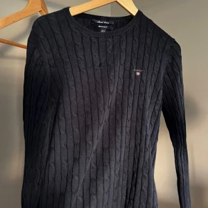 Svart kabelstickad tröja från Gant - Kabelstickad marin blå tröja från Gant med rund halsringning och diskret logga på bröstet. Tröjan har lång ärm och klassisk passform, perfekt för en stilren look. Materialet är mjukt och bekvämt, passar till många olika outfits. Lite luddigt.