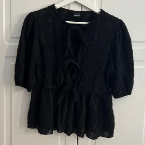 Svart blus med knyt från Gina Tricot - Supersnygg svart blus från Gina Tricot i storlek XS. Blusen har ett vågigt strukturerat tyg, trekvartsärmar och tre rosetter framtill som man kan knyta om själv. Har bara använt den 1 gång