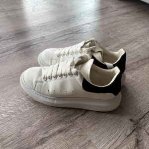 Alexander McQueen skor - Stilrena vita sneakers från Alexander McQueen med svart mockadetalj och guldfärgad logga på hälen. Skorna har tjock sula, rund tå och klassisk snörning. Perfekta för dig som gillar en clean och lyxig look. Skick 8/10. Skicka pm vid minsta fundering