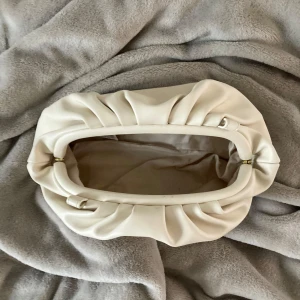 Vit kuvertväska från Even&Odd - Superfin vit kuvertväska från Even&Odd i mjukt syntetmaterial. Väskan har en cool rynkad design och avlång form, med en smal axelrem som gör den enkel att bära. Perfekt för att lyfta din outfit med en clean och modern touch. Mått på väskan i slutet.