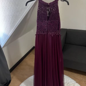 Vinröd långklänning med spets och pärlor - Magiskt snygg vinröd långklänning med spetsdetaljer och pärlor upptill. Klänningen har ärmlös design, öppen rygg och ett flowy kjolparti i tunt tyg. Perfekt för bal eller fest, med vackra broderier och elegant fall.