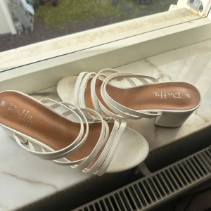 Vita sandaler med klack från Duffy - Snygga vita sandaler från Duffy med flera tunna remmar och bred klack. Skorna har öppen tå och en stilren design som passar perfekt till sommarens outfits. Tillverkade i syntetmaterial med en bekväm innersula.