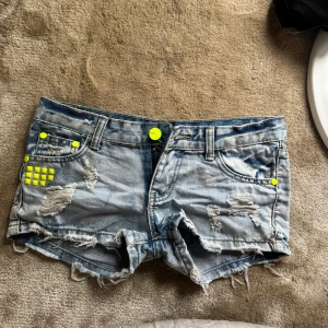 Lågmidjade Ljusa jeansshorts  - midjemått 39cm❤️Snygga ljusblå jeansshorts med slitna detaljer och fransig kant. Shortsen har neon-gula nitar och knappar som ger en cool kontrast. Klassisk femficksmodell med låg midja och råa kanter. Perfekta för sommarens alla äventyr.