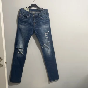 Blå slitna jeans Abercrombie & Fitch - Säljer ett par blå jeans slim fit från Abercrombie & Fitch med snygga slitningar och hål på benen. Klassisk femficksmodell med raka ben och normal passform. Jeansen har tydliga slitdetaljer framtill och är tillverkade i bomull för en skön känsla.