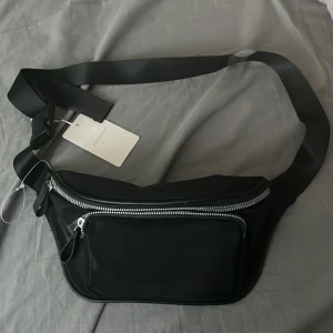 Svart axelväska från Primark - Snygg svart axelväska från Primark med bred rem och stora silvriga dragkedjor. Väskan har ett stort huvudfack och en praktisk ytterficka framtill. 