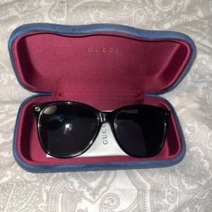 Svarta solglasögon från Gucci - Snygga svarta solglasögon från Gucci med klassisk wayfarer-form. Bågarna är i blank svart plast med diskret guldfärgad Gucci-logga vid gångjärnen. Kommer med originalfodral i röd sammet och blå kant. Perfekta för att lyfta din stil.