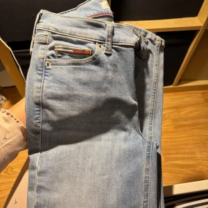 Ljusblå jeans från Tommy Jeans - Säljer ett par ljusblå jeans från Tommy Jeans med klassisk femficksdesign och broderad logga på bakfickan. Jeansen har normal passform, raka ben och är tillverkade i mjuk bomullsdenim och har låg midja. Perfekta för dig som gillar en clean och tidlös look.