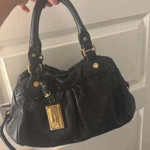 Svart handväska Marc by Marc Jacobs - Snygg svart handväska i skinn från Marc by Marc Jacobs med guldiga metalldetaljer och dragkedja. Väskan har dubbla handtag, dekorativa nitar och en ikonisk metallbricka framtill. Fodret har ett coolt mönster med märkets logga.