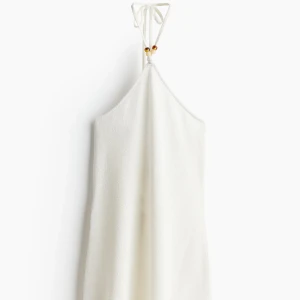 Vit kort halterneckklänning med pärlor - Superfin vit kort klänning i frotté med halterneck och knyt i nacken, dekorerad med små pärlor. Perfekt för varma dagar och ger en chill vibe. Enkel och stilren design med mjukt material som känns skönt mot huden. 