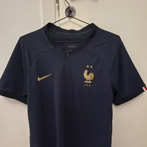 Frankrike Nike fotbollströja marinblå - Snygg marinblå fotbollströja från Nike med Frankrikes landslagsmärke och två guldstjärnor på bröstet. Guldigt Nike-märke och FFF-logga. Kortärmad modell i fint material, perfekt för match eller träning.