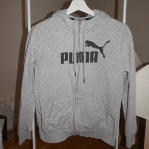 Grå hoodie från Puma med dragkedja - Grå hoodie från Puma med svart logga på bröstet. Tröjan har huva med snörning, hel dragkedja framtill och två fickor. Perfekt för chill dagar eller när du vill ha en sportig look. Mjuk och skön i bomullsmix.