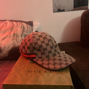 Mönstrad keps från Gucci - Snygg keps från Gucci med det klassiska GG-mönstret i beige och brunt. Kepsen har en justerbar rem baktill och detaljer i rött och grönt på sidan. Perfekt för att ge din outfit en lyxig touch.