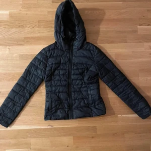 Svart quiltad jacka med huva - Snygg svart quiltad jacka med dragkedja och huva. Perfekt för kyligare dagar. Jackan har långa ärmar och en stilren design.
