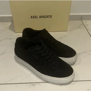 Svarta sneakers från Axel Arigato - Snygga svarta sneakers från Axel Arigato med vit sula. Perfekta för en stilren look. Skorna har snörning och är i en klassisk design som passar till många olika outfits.