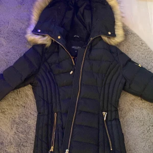 Svart dunjacka från Hollies - Säljer en snygg svart dunjacka från Hollies med pälskantad huva och dragkedja. Jackan har quiltad design och praktiska fickor med dragkedjor. Perfekt för kalla dagar!