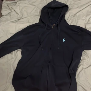 Mörkblå hoodie från Polo Ralph Lauren - Säljer en stilren mörkblå hoodie från Polo Ralph Lauren med dragkedja och en liten broderad logga på bröstet. Perfekt för en avslappnad stil. Den har långa ärmar och en praktisk huva med snörning.