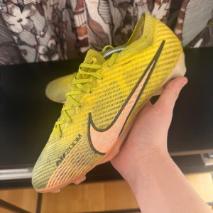 Nike mercurial - Snygga gula fotbollsskor från Nike med svart swoosh och snörning. Skorna har en modern design med Air Zoom-teknologi för extra komfort och prestanda på planen. Perfekta för den som vill sticka ut på fotbollsplanen.