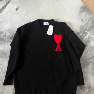 Svart tröja med rött hjärta från Ami - Säljer en stilren svart tröja från Ami med ett rött hjärta och bokstaven A på bröstet. Tröjan har långa ärmar och ribbstickade muddar vid ärmslut och nederkant. Perfekt för en trendig look.