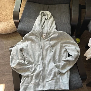 Grå hoodie från Nike - Säljer en klassisk grå hoodie från Nike med dragkedja och huva. Perfekt för en avslappnad stil. Den har långa ärmar och en liten Nike-logga på bröstet. Skön och enkel att matcha med det mesta.