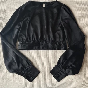 Svart satinblus - Svart blus i satin med långa ärmar och knappar vid slutet på ärmen. Blusen har en öppen rygg och är croppad. 