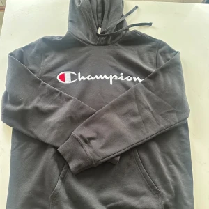 Svart hoodie från Champion - Säljer en svart hoodie från Champion med det klassiska logotyptrycket på bröstet. Tröjan har en justerbar huva med dragsko och en stor magficka framtill. Perfekt för en avslappnad stil.
